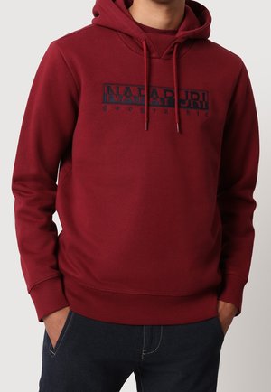Mann trägt burgunderfarbenen Kapuzenpullover mit Frontlogo „Napapijri geographic“, Hände in dunklen Jeans taschen.