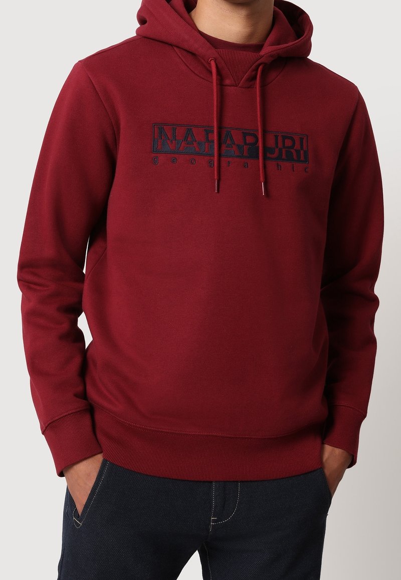 Homme portant un sweat à capuche bordeaux avec un logo devant indiquant "Napapijri geographic", mains dans les poches d'un jean foncé.