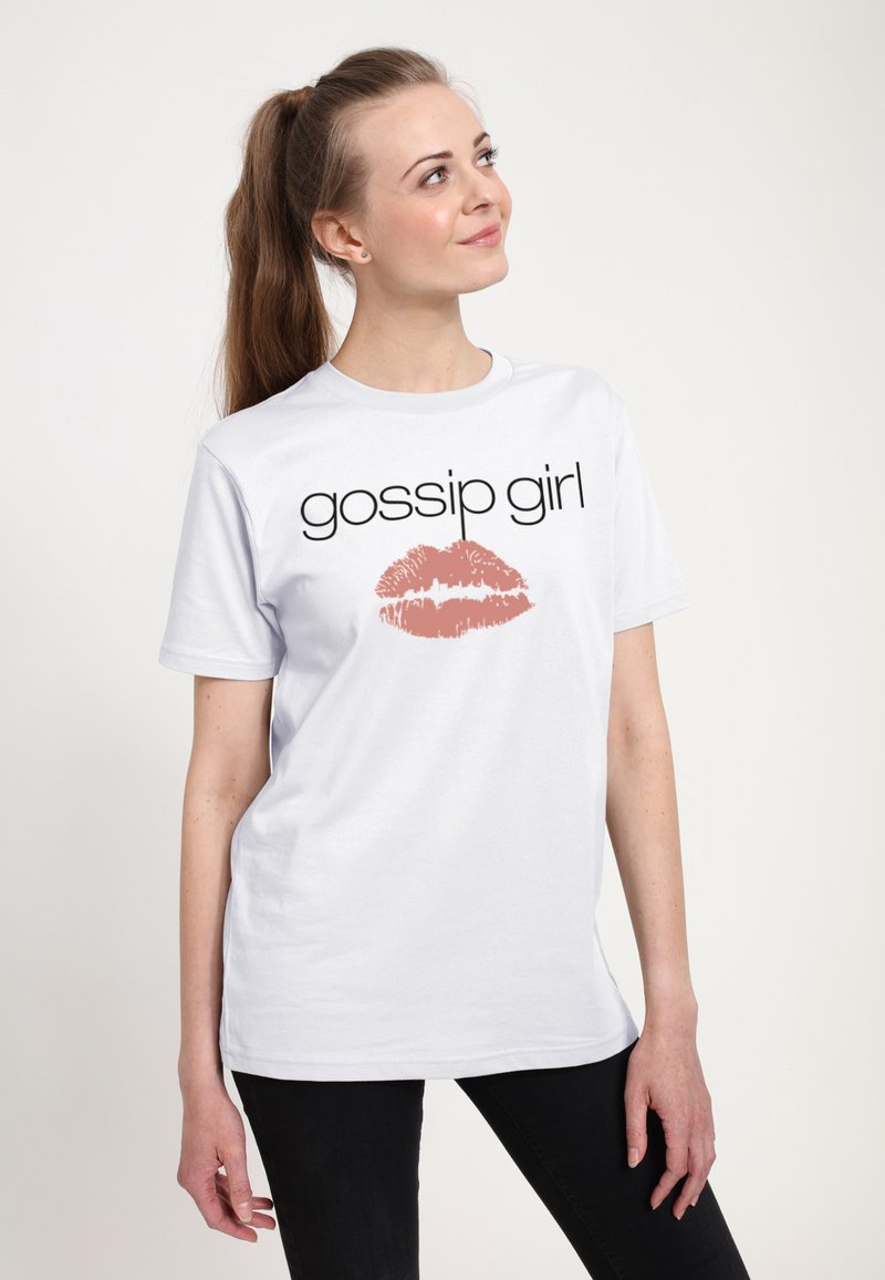 Ung kvinde med lang hestehale iført hvid "Gossip Girl" T-shirt med lyserødt læbeprint, stående foran ensartet lyst baggrund.