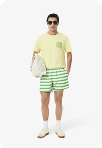 Camiseta de algodón amarilla con un bolsillo de patrón verde, combinada con pantalones cortos a rayas verdes. Calcetines blancos y zapatillas completan el conjunto.