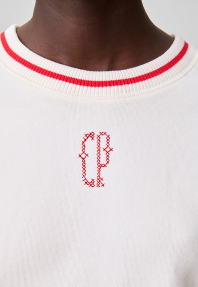 Pull blanc avec un col en tricot rouge côtelé et un logo brodé rouge sur la poitrine. Le tissu a une texture lisse et une légère élasticité.