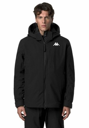 Uomo con giacca nera con cappuccio e logo bianco sul petto, in piedi dritto contro uno sfondo bianco semplice.