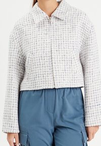 Geknipt jack van textuurwit stof met blauwe draden, voorzien van een kraag en een ritssluiting aan de voorkant. Gepaarde met losse blauwe broek.