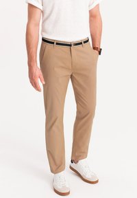 Chino beige con tasche frontali, cintura nera con hardware argentato, abbinati a sneakers bianche con suola beige e marrone.