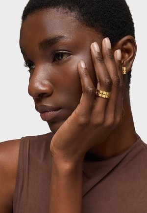 Gold jewelry on a hand, featuring a ring and earrings with a curved, scalloped design. Skin tone is deep brown, against a neutral background.