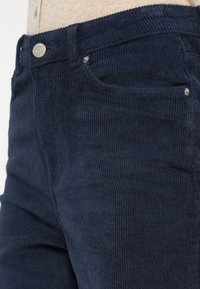 Gros plan d’un pantalon en velours côtelé bleu marine avec poche avant visible, ceinture, passants et détail du bouton métallique.