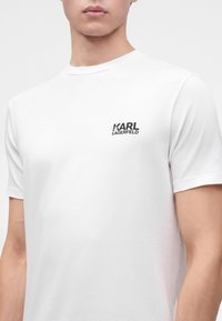 Jeune homme portant un t-shirt blanc uni avec un petit logo noir Karl Lagerfeld sur le côté gauche de la poitrine.