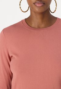 Rustfarbenes gestricktes Sweatshirt mit rundem Ausschnitt und langen Ärmeln, kombiniert mit großen, gedrehten goldenen Creolen. Glatte Textur.