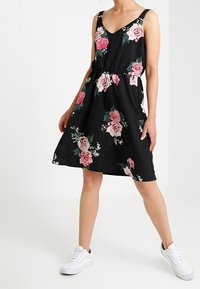 Černé květinové šaty s růžovými a bílými květinovými vzory, se vzorem do V, silnými ramínky a volným střihem, doplněné o bílé tenisky.