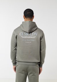 Άνδρας που φοράει γκρι φούτερ με κουκούλα και παντελόνι, γυρισμένος πλάτη, στο πίσω μέρος του φούτερ αναγράφεται το μήνυμα "CLSR LDN, Ποιοτικά ενδύματα σχεδιασμένα στο Λονδίνο."