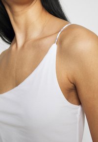Top bianco senza maniche realizzato in tessuto liscio, con un design monospalla e una sottile fascia con testo stampato lungo il bordo.