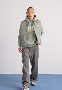 Hellgrüne Steppweste über einem passenden Kapuzenpulli, graue Cordhosen und graue Sneaker. Einfaches Design, weicher Stoff und neutrale Farben.