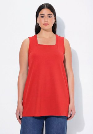 Kobieta z długimi ciemnymi włosami, nosząca złote kolczyki-koła, czerwoną, bezrękawnikową bluzkę z kwadratowym dekoltem i niebieskie dżinsy, stojąca na białym tle.