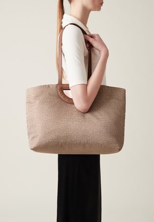 Brown patterned tote bag with textured surface and leather handles, featuring a circular cutout. Bag is held at shoulder level.