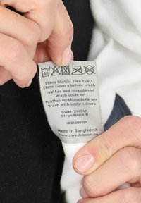 Etichetta di cura in tessuto grigio tenuta in mano, con istruzioni di lavaggio, dettagli sul prodotto e il testo "Made in Bangladesh" in basso.