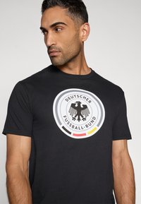 Fekete póló kör alakú logóval, amelyen a német labdarúgó címer, a "DEUTSCHER FUSSBALL-BUND" felirat, valamint piros, sárga és fekete színű díszítőelemek láthatók.