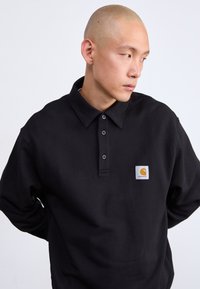 Polo shirt in cotone nero con colletto a bottoni, caratterizzata da un piccolo logo a patch quadrato in arancione e bianco. Tessuto morbido e vestibilità casual.