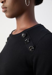 Černý svetr s dekorativními černými kameny na ramenou. Tkanina vypadá hladce a má klasický pruhovaný výstřih.