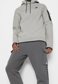 Grå Nike-hoodie med dragsko, ficka med dragkedja på ärmen och svarta detaljer. Matchas med grå joggers med cargofickor och logotyp.