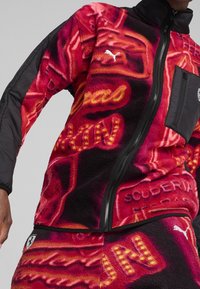 Red fleece jacket with black sleeves, featuring bold yellow and orange patterns, a zip closure, and a chest pocket with a logo.