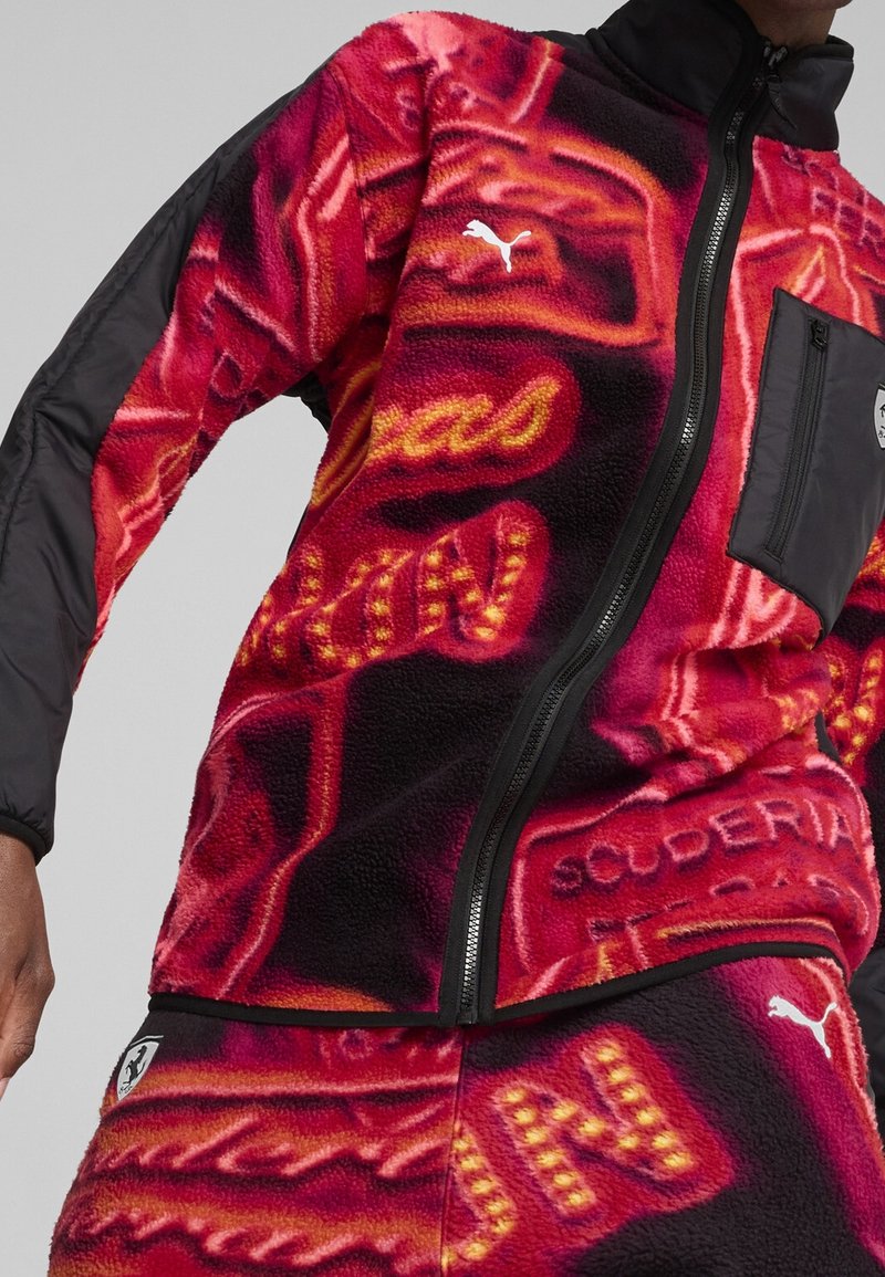 Red fleece jacket with black sleeves, featuring bold yellow and orange patterns, a zip closure, and a chest pocket with a logo.