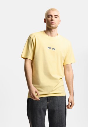 Młody mężczyzna z krótkimi blond włosami, ubrany w bladożółtą koszulkę z napisem „PROSTO.” oraz ciemne, luźne dżinsy na jednolitym tle.