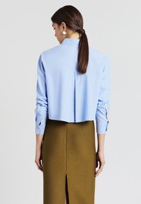 Frau mit dunklem Haar im Zopf, die ein hellblaues, langärmliges, cropped Shirt und einen olivgrünen, hoch taillierten Rock mit einem Schlitz auf der Rückseite trägt.