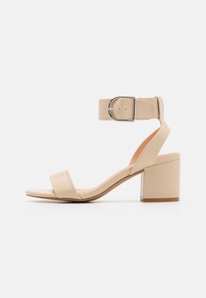 Beige offene Sandale mit Blockabsatz und breitem Knöchelriemen mit silberner Schnalle auf weißem Hintergrund.