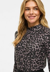 Femme souriante avec de longs cheveux blonds portant un haut à col montant et à manches longues présentant un motif léopard noir et gris.