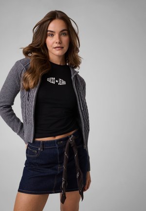 Junge Frau mit langen braunen Haaren trägt graue Strickjacke, schwarzes Guess Jeans-Shirt und einen dunklen Jeansrock mit schwarzem Schal-Gürtel, posiert vor grauem Hintergrund.