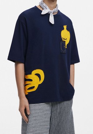 Personne portant un t-shirt oversize bleu marine avec un vase jaune et une broderie abstraite, un bandana blanc autour du cou, et un pantalon à motifs noir et blanc.