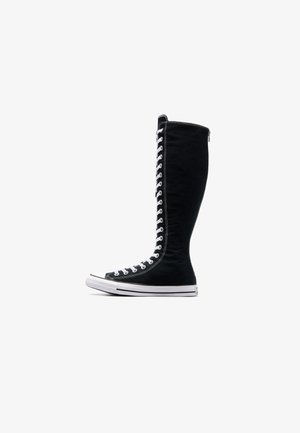 Adidași negri cu glezna înaltă, cu un design elevat, realizați din pânză. Acestia au șireturi albe, cusături și o talpă din cauciuc cu capac pentru degete.