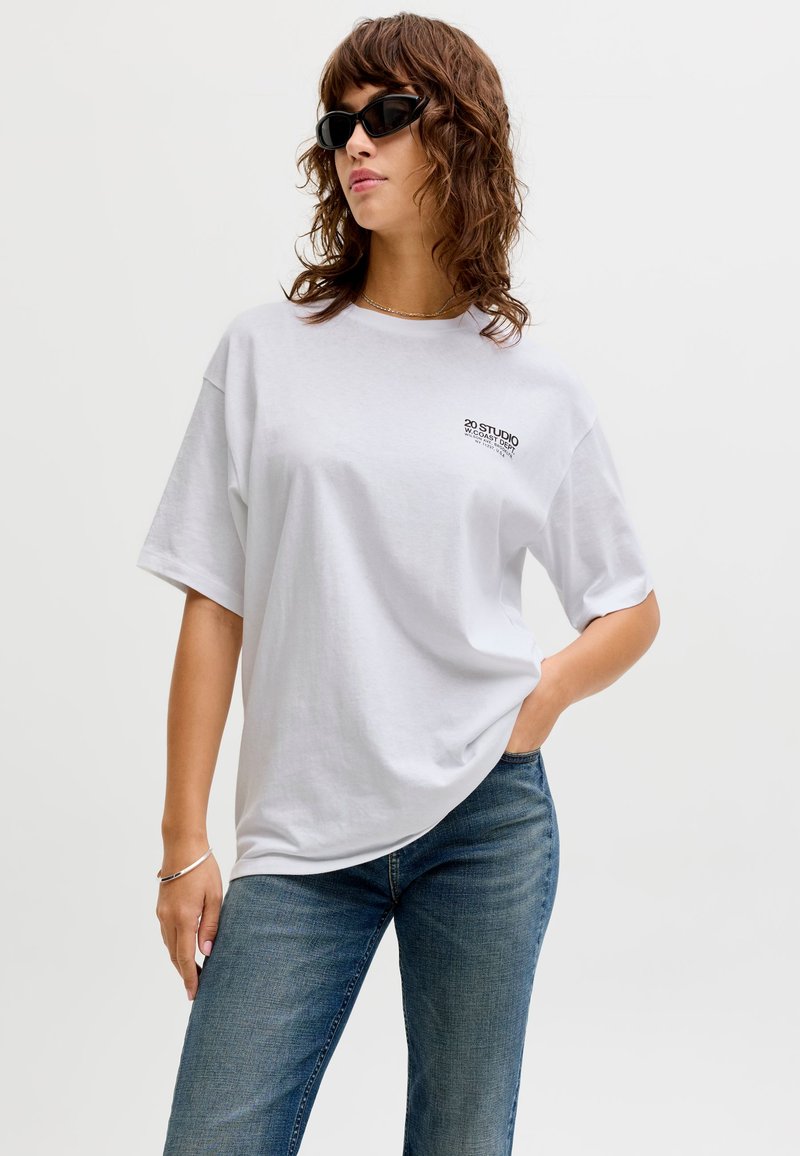 Donna che indossa occhiali da sole neri, t-shirt bianca oversize con piccolo testo sul petto sinistro, jeans blu e braccialetto d'argento al polso sinistro.