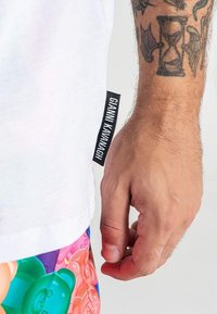 Camiseta de algodón blanca con una etiqueta negra que dice "CIANNA KAVANAGH" en el lado. Pantalones cortos coloridos con un diseño de ositos de gomita.