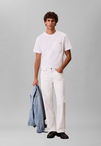 Άνδρας που φοράει άσπρο τζιν και μπλουζάκι κρατώντας ένα μπλε τζιν σακάκι, στέκεται μπροστά σε ένα απλό γκρι φόντο.