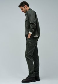 Donkergroene jas met knoopsluiting, gelaagd over een licht shirt, gecombineerd met bijpassende groene broek en bruine veterschoenen.