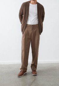 Brauner Strickcardigan über einem weißen T-Shirt, kombiniert mit hellbraunen Hosen. Braune Schuhe mit kontrastierenden weißen Sohlen. Glatter, lässiger Look.