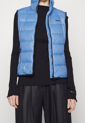 Osoba ubrana w jasno niebieską pikowaną kamizelkę bez rękawów na czarnym prążkowanym topie z długim rękawem i luźnych czarnych spodniach, stojąca na jednolitym tle.