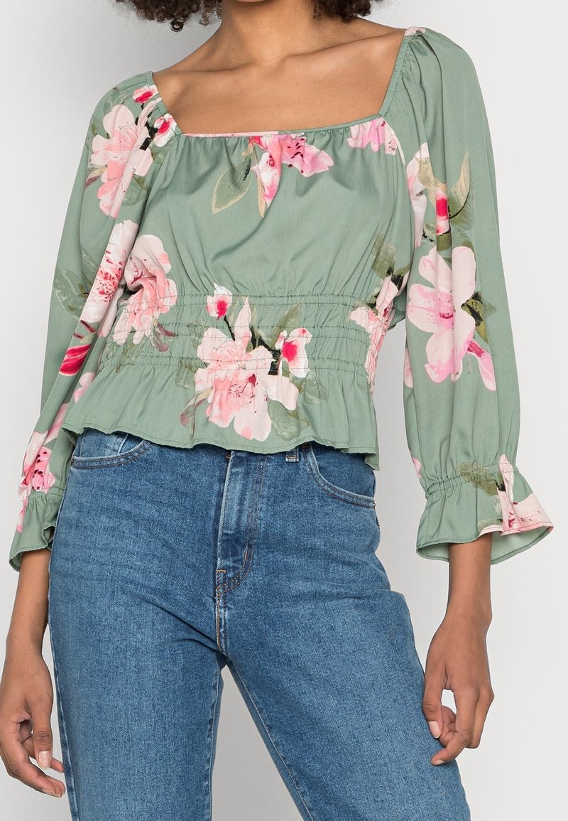Donna che indossa una blusa a maniche lunghe verde a fiori con vita arricciata e jeans blu a vita alta, in piedi contro uno sfondo neutro.