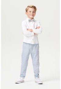 Witte langemouwen overhemd met knoopdetails, grijze vlinderdas, lichtblauwe trainingsbroek en witte sneakers met rode accenten, staat zelfverzekerd.