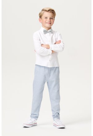 Witte langemouwen overhemd met knoopdetails, grijze vlinderdas, lichtblauwe trainingsbroek en witte sneakers met rode accenten, staat zelfverzekerd.