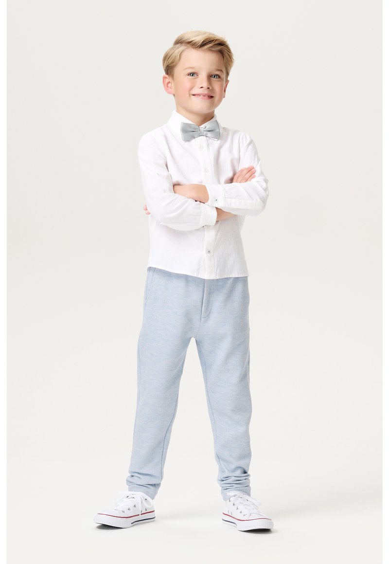 Witte langemouwen overhemd met knoopdetails, grijze vlinderdas, lichtblauwe trainingsbroek en witte sneakers met rode accenten, staat zelfverzekerd.