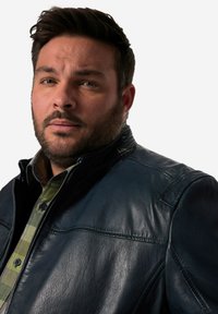 Mörkblå läderjacka med hög krage och strukturerade axlar, med en slät yta och dragkedjestängning; bärs över en grön rutad skjorta.