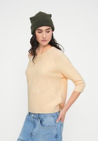 Ανοιχτό ροδακινί ριμπ πουλόβερ με φαρδιά λαιμόκοψη και μανίκια τριών τετάρτων, σετ με ψηλόμεσα μπλε τζιν σορτς και πράσινο πλεκτό σκουφάκι.