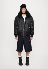 Zwarte leren bomberjack met een bontkraag, wit shirt, donkere denim cargo shorts en zwarte enkelboots. Model staat tegen een witte achtergrond.