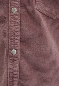 Kordstoff in Mauve mit vertikaler Rippstruktur, ausgestattet mit Metall-Druckknöpfen entlang der Knopfleiste und einer aufgesetzten Tasche auf der linken Seite.