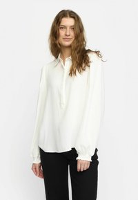 Weiße Bluse mit langen Ärmeln, Kragen, Knopfleiste und elastischen Bündchen. Hergestellt aus glattem Stoff mit lockerer Passform. Es werden schwarze Hosen getragen.