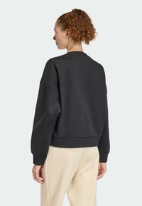 Woman with curly hair in a black long-sleeve sweatshirt and beige pants, facing away, against a plain light background.