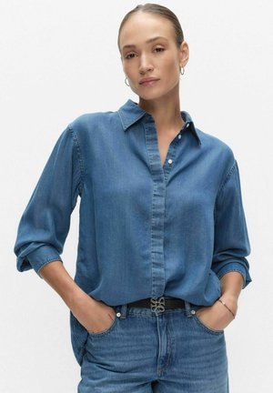 Kvinde med bagstrøget hår iført store hoop-øreringe, en blå denimskjorte med op rullede ærmer og blå jeans med sort bælte, hænderne i lommerne.