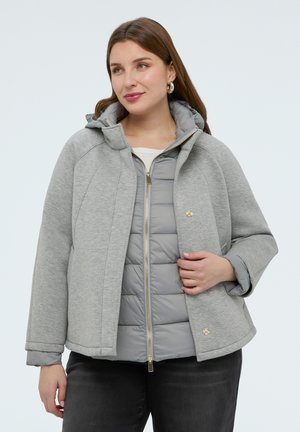 Kobieta ubrana w warstwowe szare kurtki, jedna pikowana z zamkiem błyskawicznym, a zewnętrzna z zatrzaskami, w połączeniu z czarnymi spodniami.